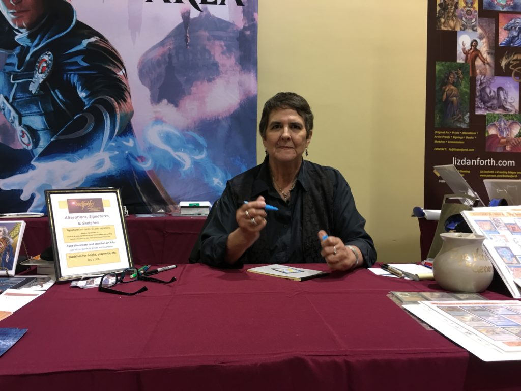 Dark-haired woman Liz Danforth, wearing a black shirt, holding a pen and preparing to sign a card in front of her on the table at a convention.